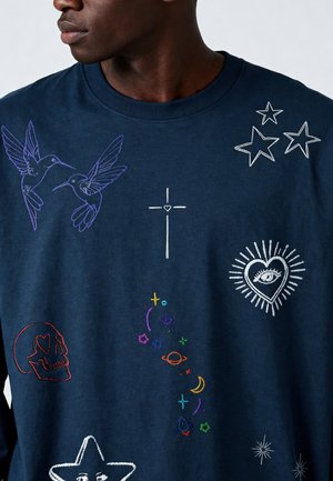Uomo che indossa una felpa blu navy con motivi ricamati colorati di colibrì, stelle, croce, occhio nel cuore, teschio e simboli celesti.