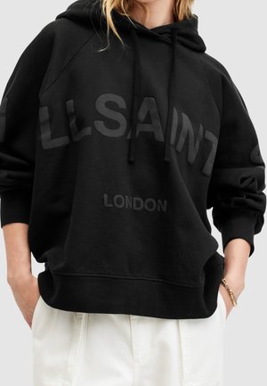 Henkilö, jolla on päällään musta huppari, jossa on edessä teksti "ALLSAINTS LONDON", yhdistettynä valkoisiin housuihin ja kultaan rannekoruun.