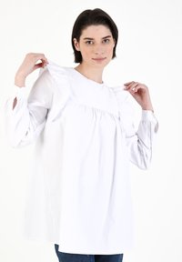 Weißes Bluse mit langen Ärmeln und hohem Hals, Rüschendetails an den Schultern und leicht gerafftem Brustbereich, mit geknöpften Manschetten.