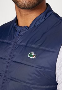 Colete acolchoado azul-marinho feito de um tecido brilhante e suave, com fecho completo e um pequeno logótipo de crocodilo verde bordado no peito.