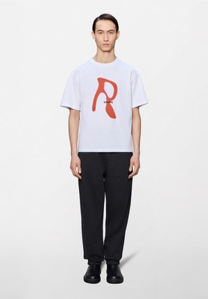 Man with long dark hair wears white t-shirt with large red letter "R" and numbers, black pants, and black shoes, standing against white background.