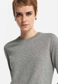 Szary sweter z dzianiny o okrągłym dekolcie, gładkiej teksturze i długich rękawach, noszony przez modelkę z krótkimi włosami i kolczykami wkrętami.