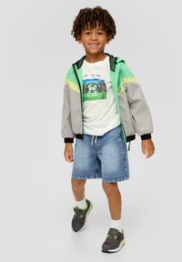 Giacca bomber grigia e verde, t-shirt bianca con stampa di un pallone da calcio, pantaloni corti di jeans e sneakers grigie con accenti verdi. Bambino sorridente in posa.