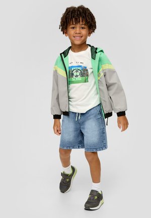 Giacca bomber grigia e verde, t-shirt bianca con stampa di un pallone da calcio, pantaloni corti di jeans e sneakers grigie con accenti verdi. Bambino sorridente in posa.