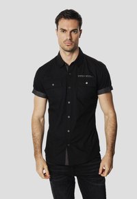 Camisa negra de manga corta con botones, dos bolsillos en el pecho, herrajes plateados y mangas remangadas. Hecha de una tela suave y ligera.