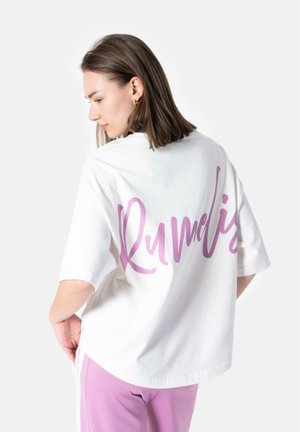 Bílý oversized tričko s fialovým kurzívním textem "RUMELIS" na zadní straně. Model ho kombinuje s růžovými kalhotami, které mají uvolněný střih a bavlněnou texturu.