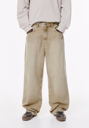 Beige wide-leg jeans with a high waist, two side pockets, and faded texture, paired with gray and brown sneakers.