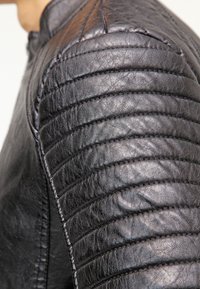 Giacca in pelle nera con dettagli di cuciture trapuntate sulle spalle, caratterizzata da coste verticali e una texture liscia. Chiusura con zip non visibile.