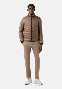 Lichtbruine pufferjas met een hoge kraag, verticale stiksels en zakken met rits, gecombineerd met beige broek en witte sneakers.