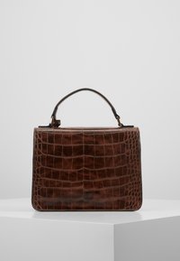Sac en cuir marron avec motif de crocodile, doté d'une poignée supérieure unique, présenté sur un piédestal blanc contre un fond gris uni.