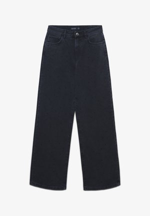 Jeans large à jambes larges de couleur noire avec une texture à pois subtile, boutons avant, fermeture éclair et design à cinq poches, étendus à plat sur un fond blanc.