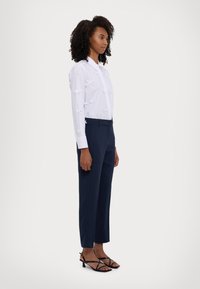 Chemise blanche à boutons avec des accents de nœuds texturés, associée à un pantalon taille haute navy et des talons noirs à lanières, présentée en vue de côté.