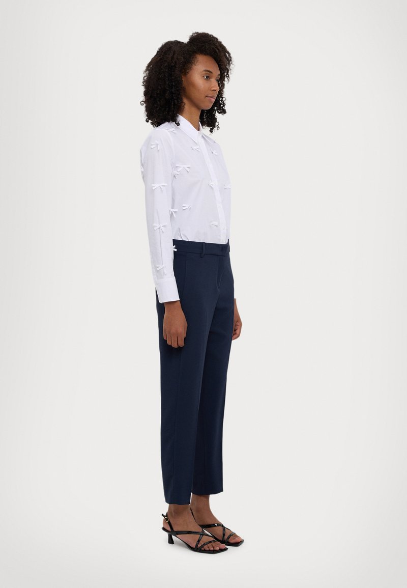 Chemise blanche à boutons avec des accents de nœuds texturés, associée à un pantalon taille haute navy et des talons noirs à lanières, présentée en vue de côté.