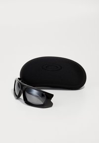 Óculos de sol envolventes pretos com lentes escuras colocados à frente de uma caixa rígida preta com fecho éclair sobre uma superfície branca.
