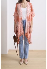 Lichtgewicht koraalkleurige kimono met witte ingewikkelde patronen en franjes aan de zoom, gecombineerd met blauwe jeans en naakte puntige platte schoenen.