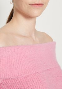 Maglione rosa off-the-shoulder, texture lavorata a maglia con motivi a coste orizzontali, caratterizzato da una finitura morbida e pelosa.