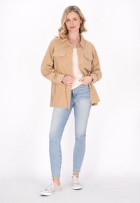 Beige fløjlskjortjakke med to brystlommer, knaplukning og en afslappet pasform, parret med lyseblå skinny jeans og hvide sneakers.