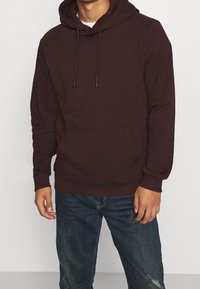 Sudadera color marrón con cordones, bolsillo tipo canguro, puños y dobladillo acanalados, hecha de tela suave. Combinada con jeans de mezclilla desgastados.