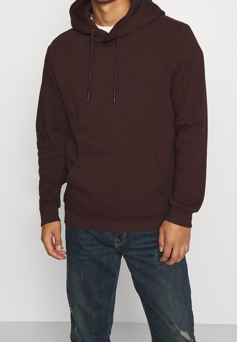 Sudadera color marrón con cordones, bolsillo tipo canguro, puños y dobladillo acanalados, hecha de tela suave. Combinada con jeans de mezclilla desgastados.