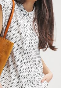 Kvinna med långt brunt hår som bär en vit skjorta med svarta rutor, bärande en senapsgul mockaväska, hand i fickan.