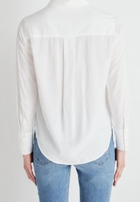 Chemise blanche à manches longues avec un col, présentant un détail de pli au dos. Elle a des poignets à boutons et une texture lisse et légère. Portée avec un jean bleu clair.