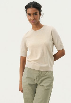 Un pull en tricot beige à manches courtes avec un col rond, fabriqué dans un matériau doux, associé à un pantalon vert clair. Texture lisse, design minimal.