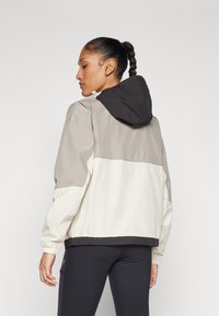 Chaqueta ligera en bloques de color con capucha negra, paneles en beige y gris, puños y dobladillo elásticos, y una textura suave.