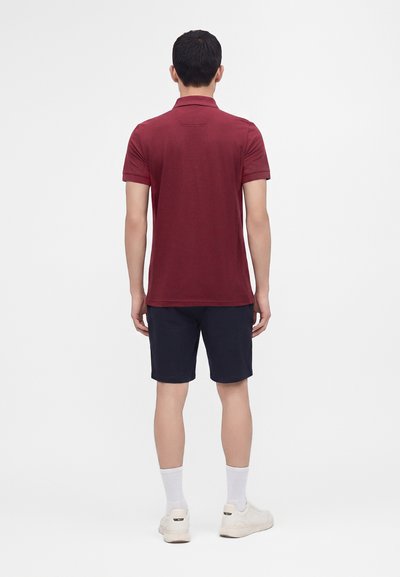Homme debout de dos portant un polo à manches courtes bordeaux, un short marine, des chaussettes blanches et des baskets blanches sur un fond uni.