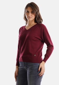 Donna con capelli castani mossi che indossa un maglione bordeaux con scollo a V e jeans blu, in piedi con una mano sulla vita davanti a uno sfondo semplice.