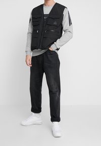 Svart multifickväst med dragkedja, bärs över en grå sweatshirt och matchas med mörka jeans och vita sneakers.