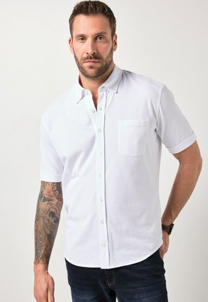 Mężczyzna z brodą w białej, krótkim rękawem koszuli na guziki, ciemnych dżinsach i zegarku na nadgarstku, pokazujący szczegółowy tatuaż czaszki na lewym przedramieniu.