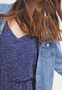 Veste en denim bleu sur une robe texturée bleu foncé avec un décolleté en V. Mettez l'accent sur le décolleté et les boutons de la veste. Cheveux légèrement ébouriffés.