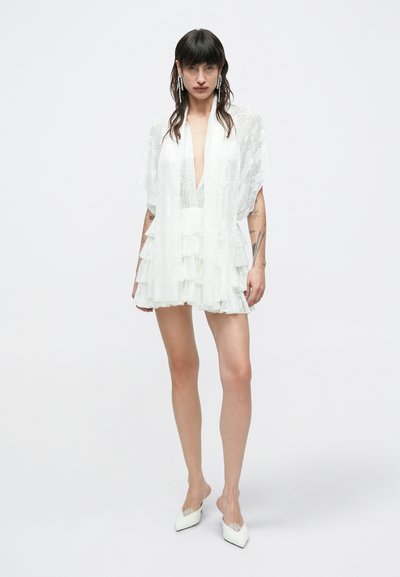 Biele vrstvené mini šaty s hlbokým výstrihom do V a priehľadnou kvetinovou látkou. Disponujú voľnými rukávmi a mäkkou textúrou. Doplnkom sú biele špicaté topánky.