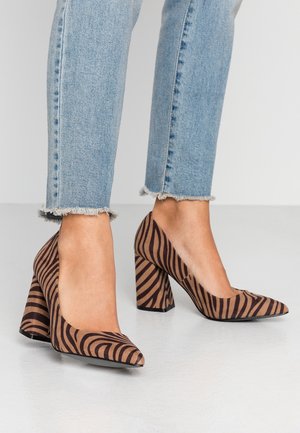 Chaussures à talons hauts à rayures marron et beige avec un bout pointu et un large talon bloc, fabriquées en tissu texturé, associées à un jean bleu effiloché.
