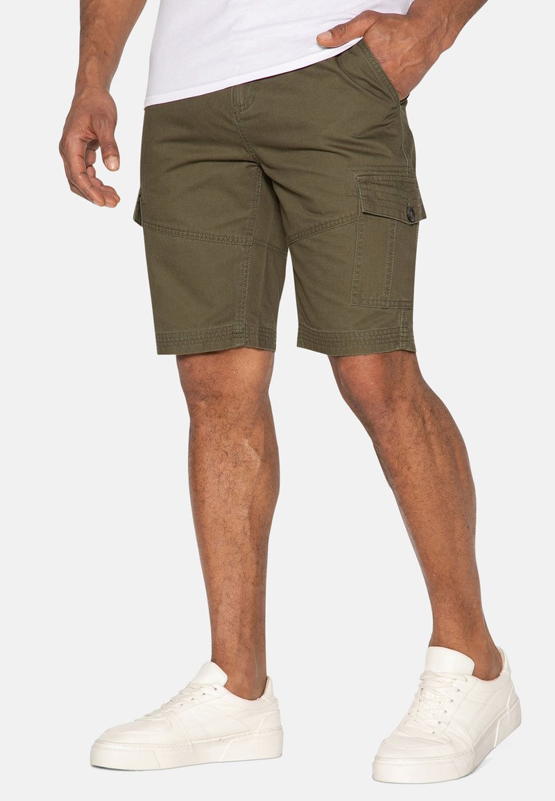 Pantalones cortos cargo de color verde oliva, confeccionados con una mezcla de algodón, que cuentan con múltiples bolsillos, costuras reforzadas y un corte recto. Zapatillas blancas completan el conjunto.