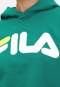 Felpa verde con grande logo bianco "FILA" e accento giallo, che mostra un primo piano del tessuto e del design del logo.
