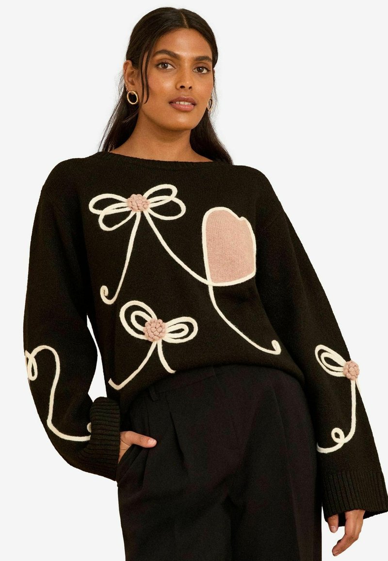 Maglione nero con ricami bianchi a forma di fiocchi e un cuore rosa. Il design include dettagli testurizzati e maniche larghe.