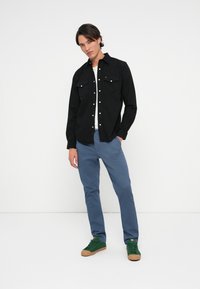 Cămașă neagră cu nasturi, cu două buzunare la piept, asociată cu pantaloni albaștri și adidași verzi cu tălpi bej. Design minimalist, croială casual.