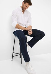 Witte button-up shirt, donkerblauwe jeans en witte sneakers. Model zit op een zwarte kruk, toont een relaxte fit en opgerolde mouwen.