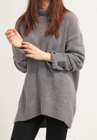 Szary, oversize sweter wykonany z miękkiego dzianiny, z wysokim kołnierzem, ściągaczami przy mankietach oraz dłuższym tylnym brzegiem.