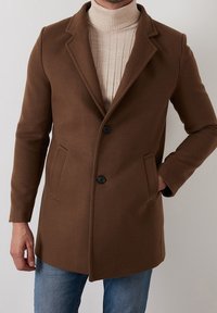 Brun ullblandad kappa med slag, två framfickor och svarta knappar. Buren över en ribbad beige polotröja, tillsammans med denimjeans.