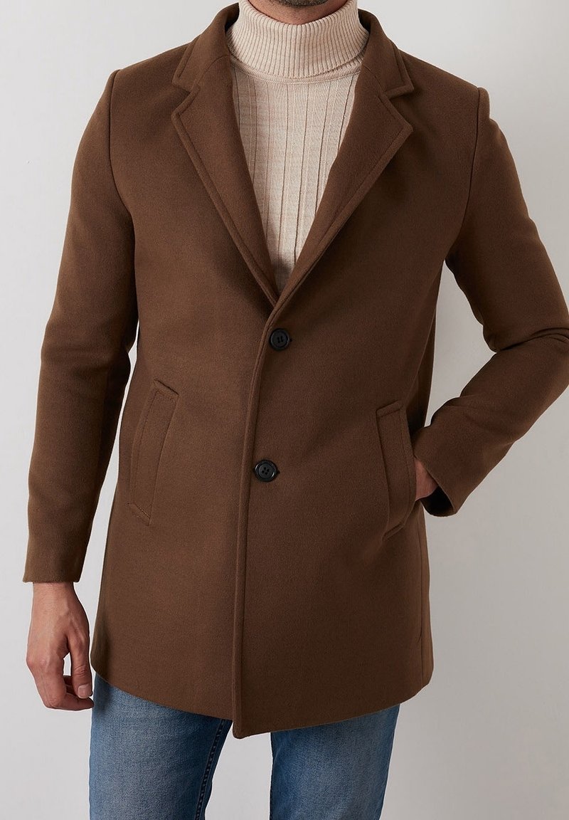 Brun ullblandad kappa med slag, två framfickor och svarta knappar. Buren över en ribbad beige polotröja, tillsammans med denimjeans.