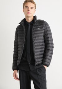 Giacca nera imbottita con trapuntatura orizzontale, colletto alto e chiusura con zip. Presenta una piccola patch con logo sul lato sinistro del petto.