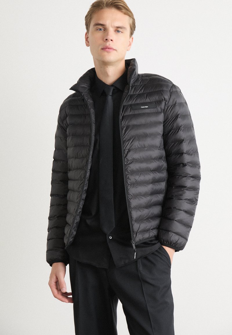 Giacca nera imbottita con trapuntatura orizzontale, colletto alto e chiusura con zip. Presenta una piccola patch con logo sul lato sinistro del petto.