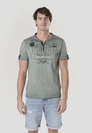 Camisa verde de manga corta con escote abotonado, con texto impreso y parches. Combinada con pantalones cortos vaqueros desgastados de color azul claro.