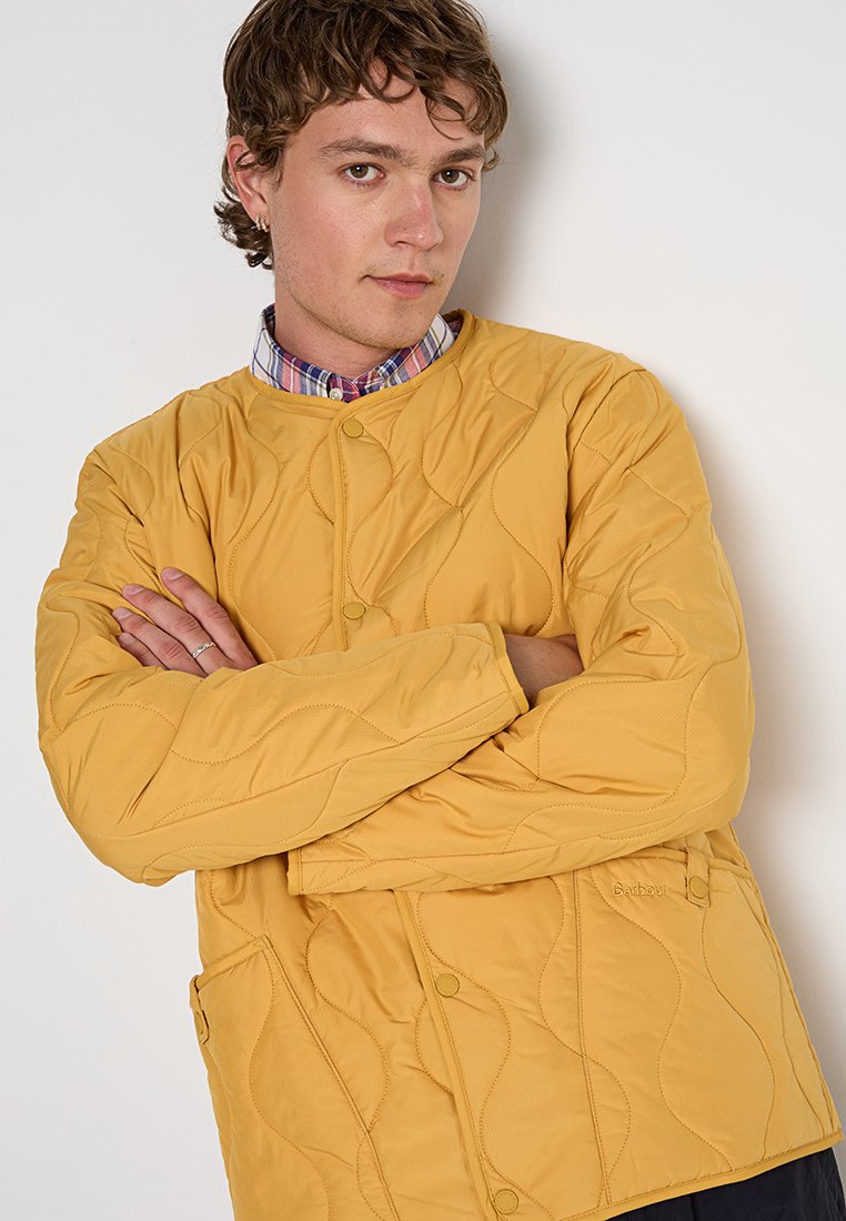 Gepolsterte gelbe Jacke mit weicher Textur, rundem Ausschnitt, Druckknöpfen und Seitentaschen. Geschichtetes Design mit sichtbaren Nähdetails.