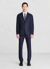 Veste de costume bleu marine avec revers crantés et deux boutons, assortie d'un pantalon coordonné. Chemise blanche et cravate verte, portées avec des chaussures de ville noires.