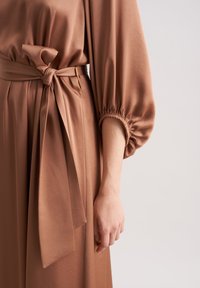 Braunes Kleid mit tailliertem Oberteil, lockeren Puffärmeln und einem gebundenen Taillengürtel. Das Material hat eine glatte Textur und fällt elegant.