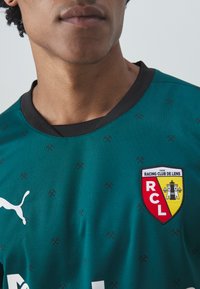 Teal fotbollströja med svart krage, strukturerad tyg och en Racing Club de Lens-logotyp i röd och gul med en slotsdesign.