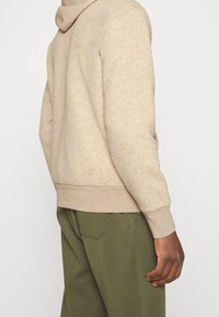 Béžová mikina s kapucí, vyrobená z měkkého materiálu, s žebrovanými manžetami a lemem, kombinovaná s olivově zelenými kalhotami, které mají uvolněný střih.